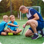 A soccer coach speaks to two young players sitting on the field.